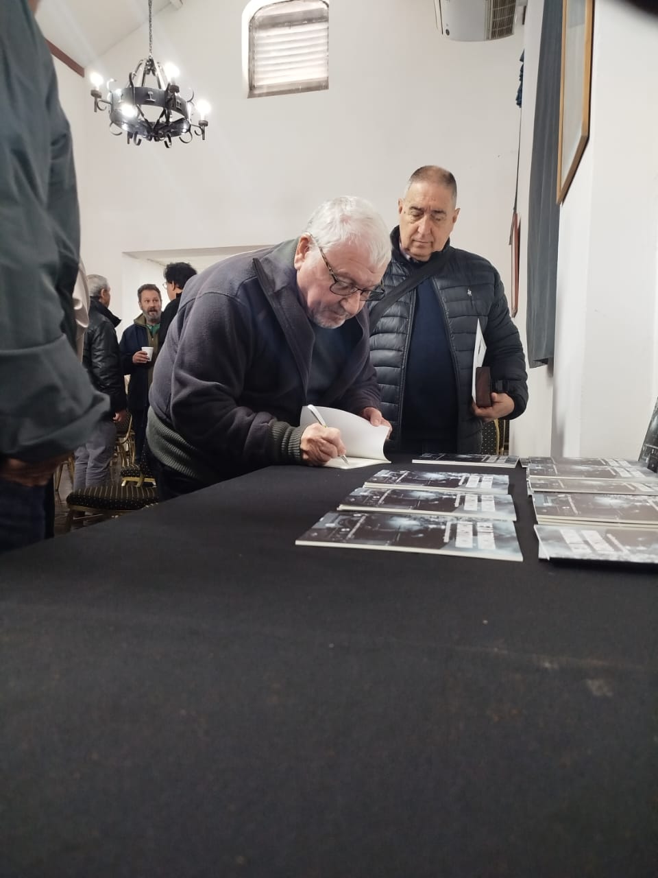 Un asistente firma ejemplares de un libro sobre una mesa, con otro asistente a su lado, en un salón con público de fondo.
